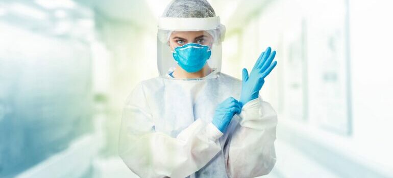Zdravstveni radnik u zaštitnoj opremi namešta plave rukavice u svetlom bolničkom hodniku.