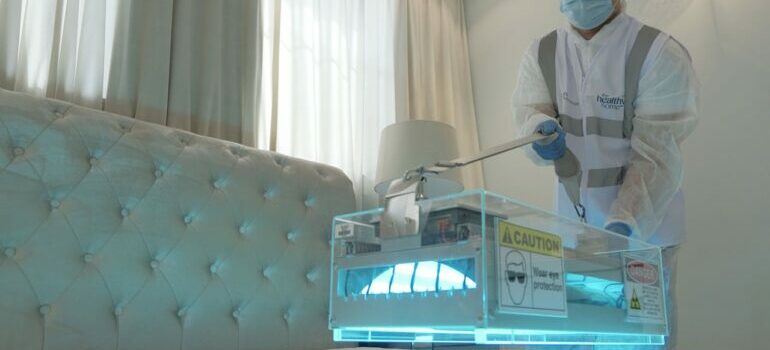 Tehničar u zaštitnoj opremi koristi UV uređaj za dezinfekciju dušeka u svetloj, modernoj spavaćoj sobi.